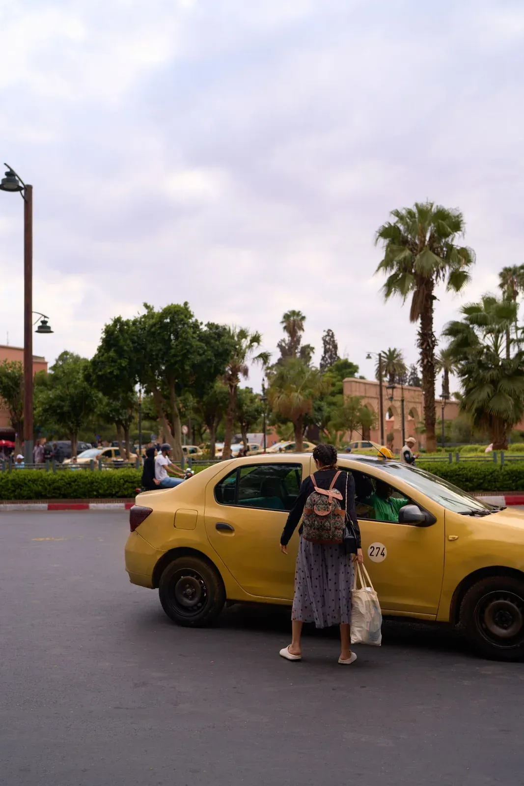 A passenger approaching a yellow taxi on a palm-lined Marrakesh street